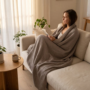 Woman reading on a sofa wrapped in a Waffle Weave Cotton Blanket in Stone